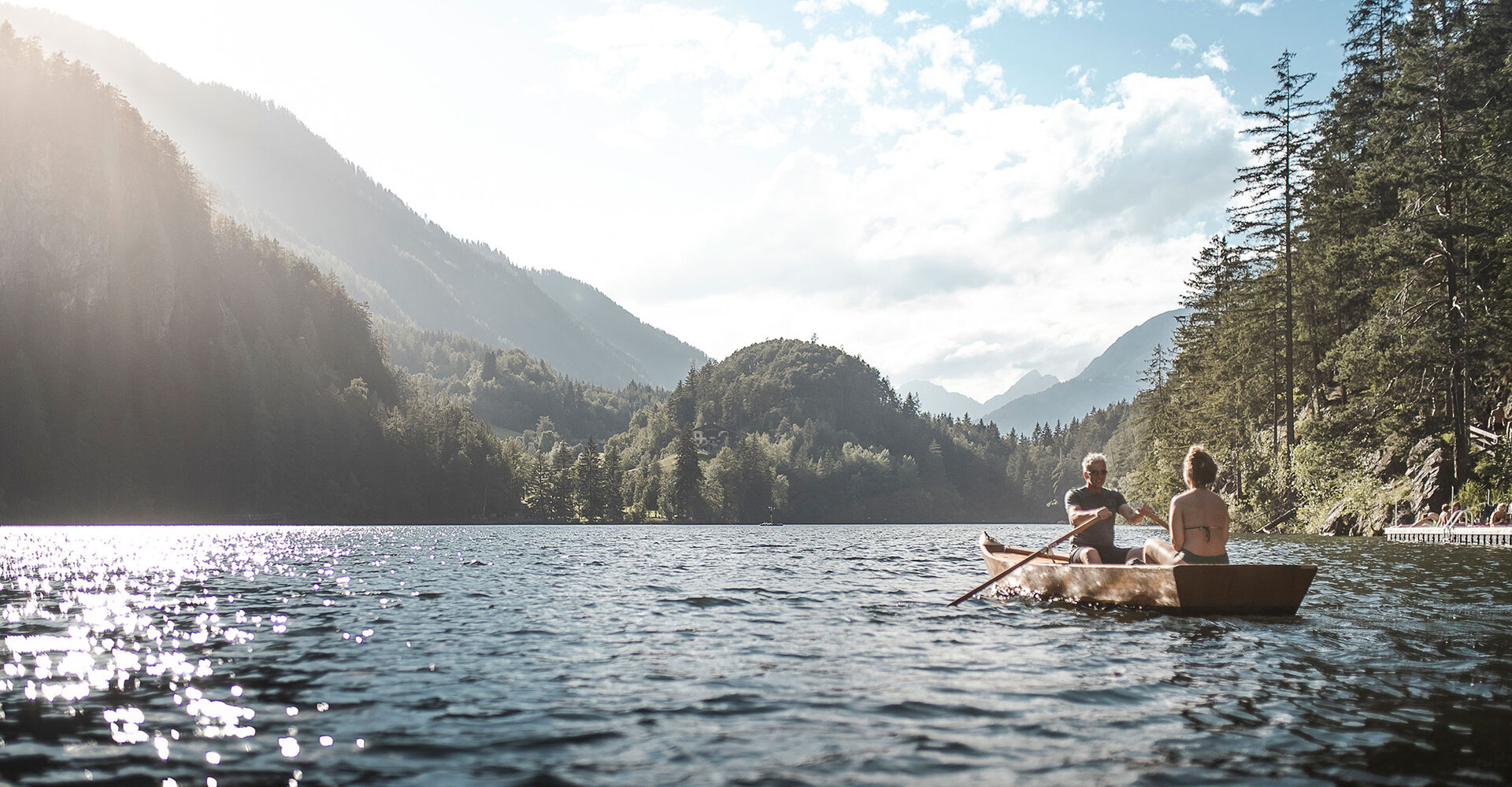 Zwei Menschen rudern in einem kleinen Holzboot auf einem ruhigen Bergsee, der von bewaldeten Hügeln und fernen Berggipfeln umgeben ist, unter einem teilweise bewölkten Himmel.