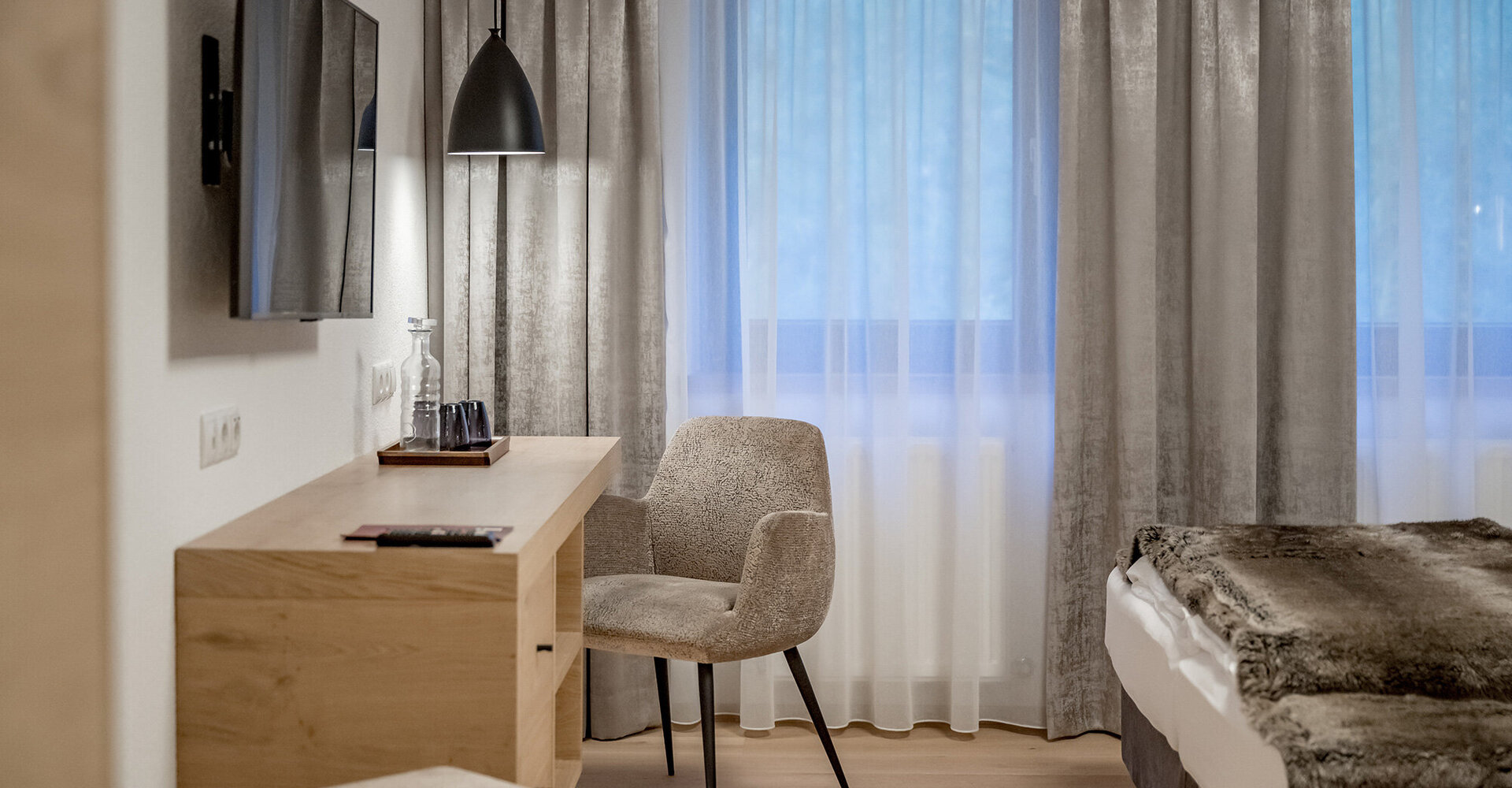 Modernes Hotelzimmer mit einem Schreibtisch aus Holz, einem beigen Stuhl, einem Flachbildfernseher, einer Hängeleuchte und einem Bett mit einer Felldecke, direkt neben großen Fenstern mit transparenten und grauen Vorhängen.