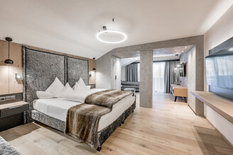 Modern hotel room with a large bed, a fur throw, nightstands, wall lamps, a TV, wooden floors, a seating area, a desk and a large window with sheer curtains allowing daylight to stream in.