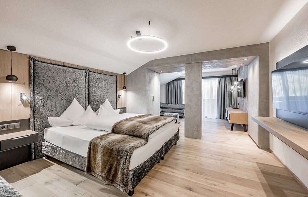 Modern hotel room with a large bed, a fur throw, nightstands, wall lamps, a TV, wooden floors, a seating area, a desk and a large window with sheer curtains allowing daylight to stream in.