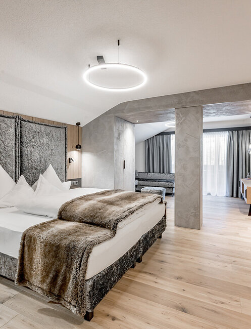Modernes Hotelzimmer mit einem großen Bett, Felldecke, Nachttischen, Wandleuchten, Fernseher, Holzböden, einer Sitzecke, einem Schreibtisch und einem großen Fenster mit transparenten Vorhängen, durch die Tageslicht hereinströmt.