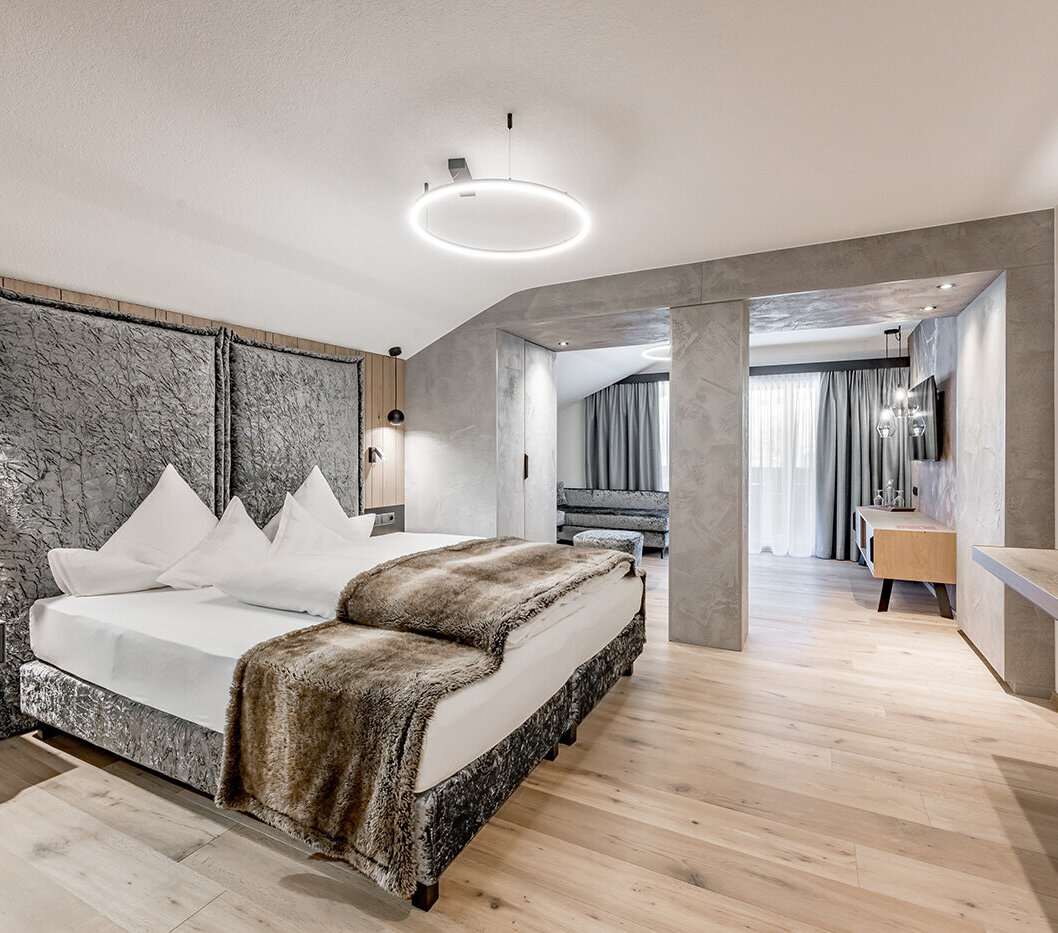 Modern hotel room with a large bed, a fur throw, nightstands, wall lamps, a TV, wooden floors, a seating area, a desk and a large window with sheer curtains allowing daylight to stream in.
