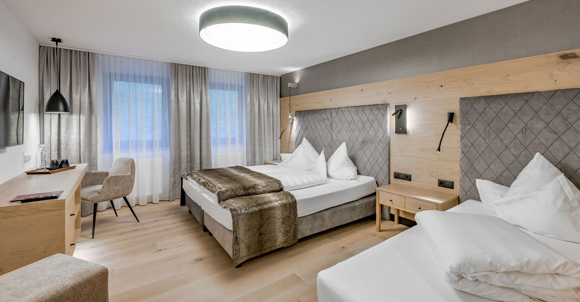 Modern hotel room with two beds, wooden flooring, gray and beige décor, a large window with curtains, a desk with a chair, a wall-mounted TV and bedside lamps.