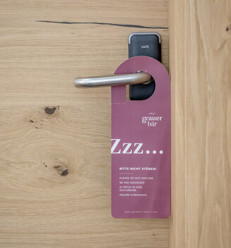 A purple door hanger with the inscription "Please do not disturb" hangs from a modern silver door handle on a light-colored wooden door.