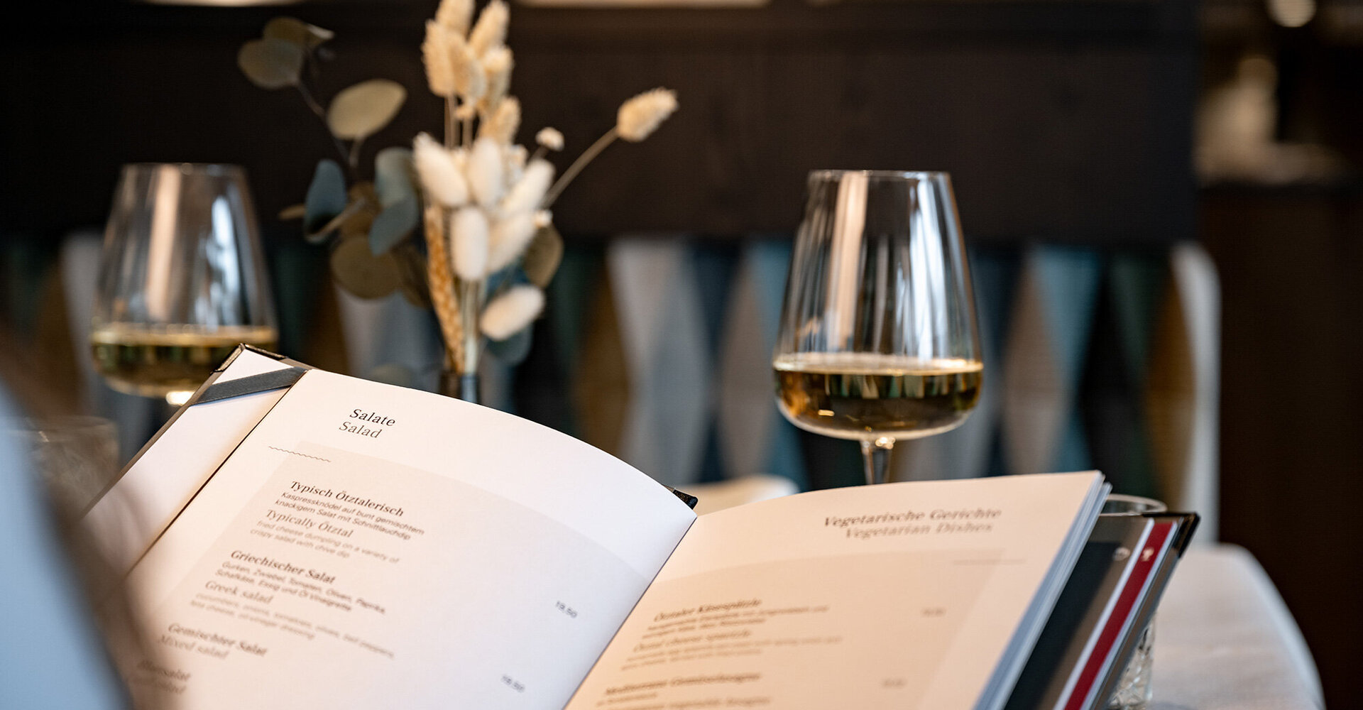 A close-up of an open menu on a table, on which there are two glasses of white wine and dried flowers in a vase, against a gently blurred background.