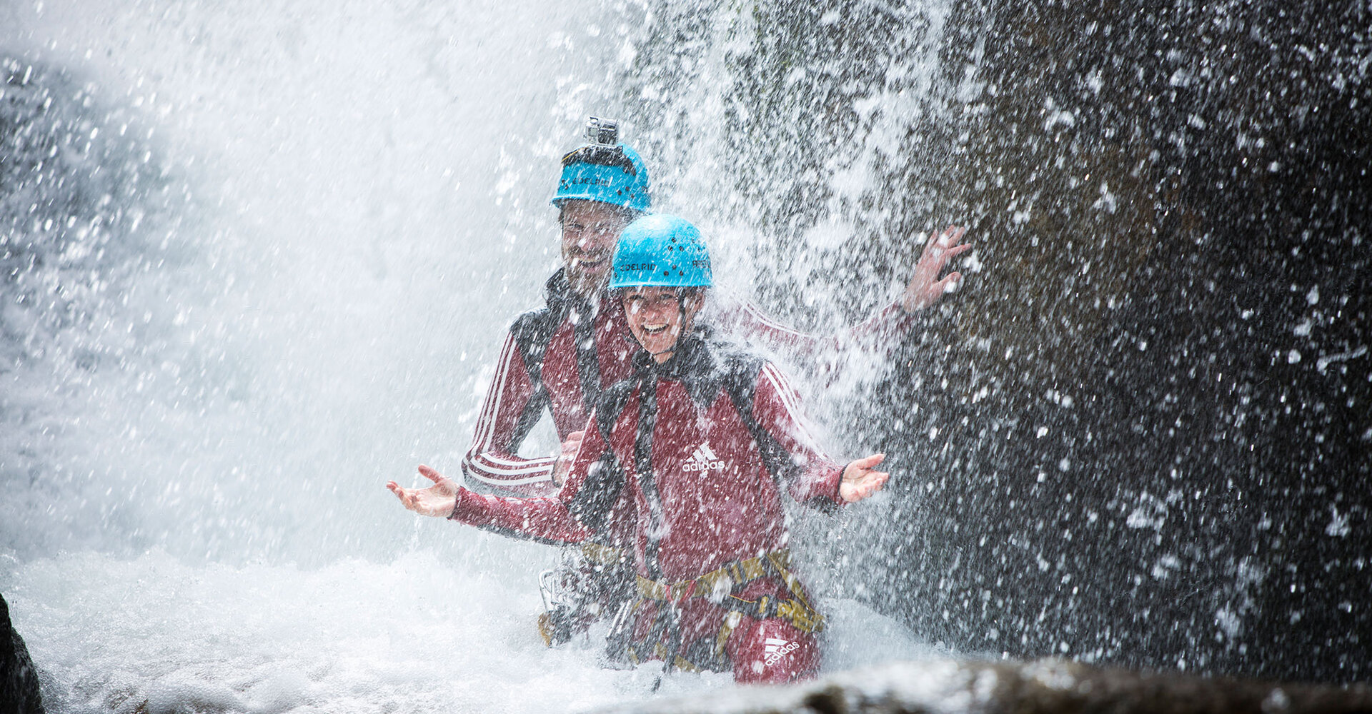 Zwei Menschen mit blauen Helmen und roten Anzügen lächeln und posieren, während sie unter einem mächtigen, sprudelnden Wasserfall stehen, umgeben von rauschendem Wasser.