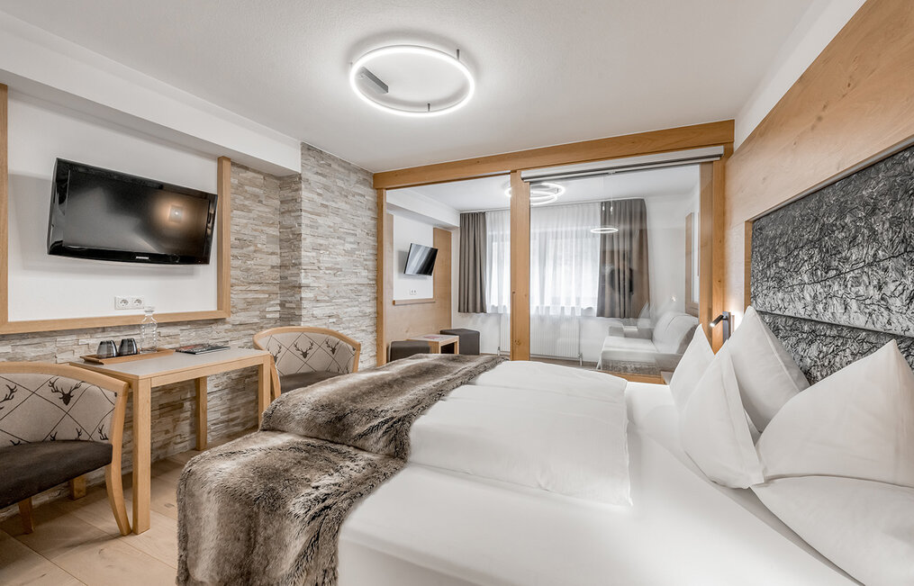 Modernes Hotelzimmer mit einem großen Bett, einer Felldecke, einem an der Wand montierten Fernseher, einem kleinen Tisch mit zwei Stühlen und einer Glastrennwand, hinter der sich eine Sitzecke mit Tageslicht befindet.
