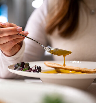 A person wearing a white turtleneck sweater holds a spoon containing yellow soup over a white bowl. A crusty loaf of bread and microgreens lie beside it.