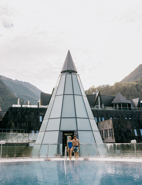 Two people in swimwear are standing at the edge of a round outdoor pool. A modern, cone-shaped glass building and mountains are in the background.