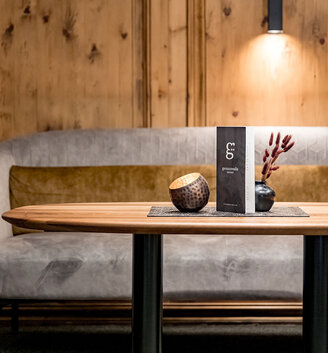 A modern lounge featuring a wooden table, a gray sofa, light brown cushions, a decorative bowl, a vase with red branches and a menu on the table, set against a wooden wall under soft overhead lighting.