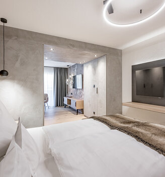 A modern hotel room featuring a neatly made bed, a faux-fur throw, a wall-mounted TV, a pendant light and a view of a bright seating area with a desk and a window. Neutral tones and minimalist décor.