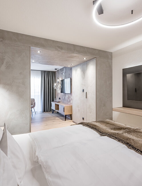 Ein modernes Hotelzimmer mit einem ordentlich gemachten Bett, einer Felldecke, einem an der Wand montierten Fernseher, einer Hängelampe und Blick auf eine helle Sitzecke mit einem Schreibtisch und einem Fenster. Neutrale Farbtöne und minimalistische Einrichtung.