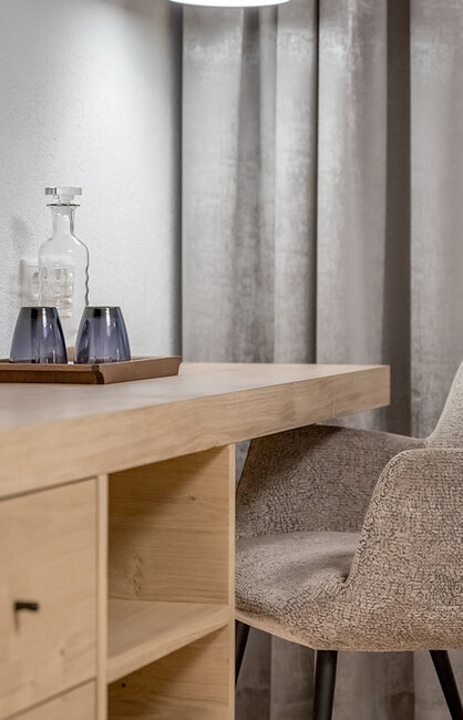 Moderne Schreibtischgarnitur in einem Hotelzimmer mit einem gepolsterten Stuhl, einem Holzschreibtisch, einem Tablett mit einer Glaskaraffe und zwei Gläsern, grauen Vorhängen, weißen Gardinen sowie einem Teil eines an der Wand montierten Fernsehers.