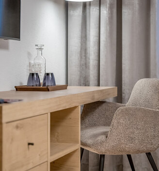 Modern desk setup in a hotel room featuring an upholstered chair, a wooden desk, a tray with a glass carafe and two glasses, gray drapes, white sheer curtains and a wall-mounted TV set.
