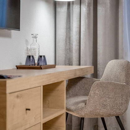 Modern desk setup in a hotel room featuring an upholstered chair, a wooden desk, a tray with a glass carafe and two glasses, gray drapes, white sheer curtains and a wall-mounted TV set.