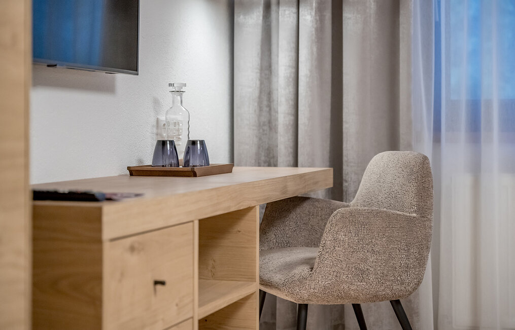 Modern desk setup in a hotel room featuring an upholstered chair, a wooden desk, a tray with a glass carafe and two glasses, gray drapes, white sheer curtains and a wall-mounted TV set.