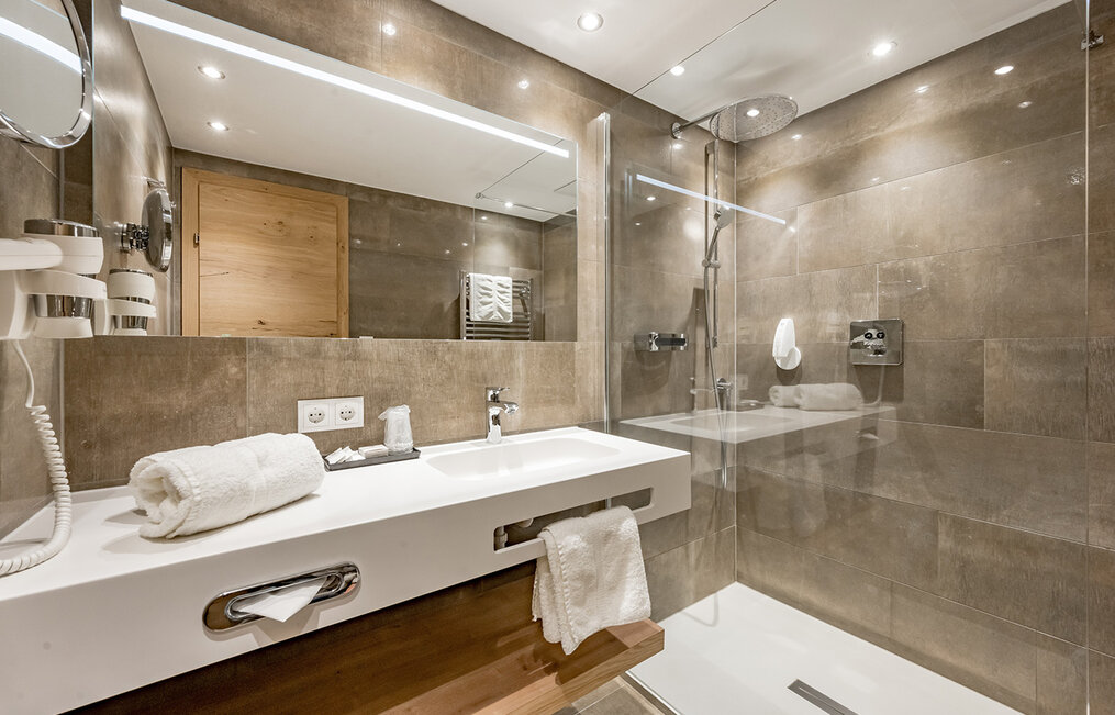 Modern bathroom with a large mirror, white sink, towel rack, walk-in glass shower, wall-mounted rainfall showerhead and light brown tiled walls under recessed lighting.