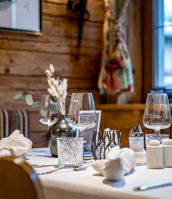 A close-up of an elegant restaurant table set with wine glasses, a vase of dried flowers, napkins and spices; wooden walls and soft lighting in the background.