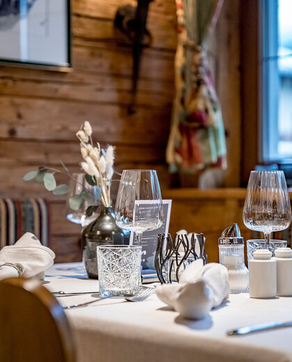 A close-up of an elegant restaurant table set with wine glasses, a vase of dried flowers, napkins and spices; wooden walls and soft lighting in the background.