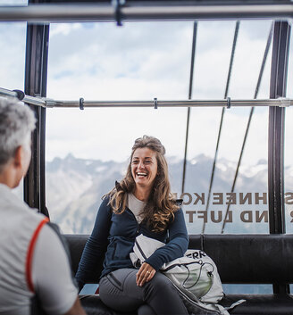 Two people are sitting in a gondola and talking, mountains are outside the windows. The woman is smiling and holding a bag, while the man sits opposite her. The interior is bright and cozy.
