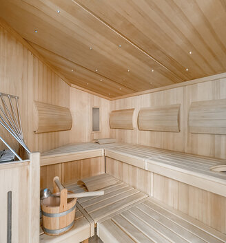 A modern wooden sauna featuring light-wood benches, walls and ceiling, equipped with a bucket and ladle, a heater with stones and curved backrests along the walls.