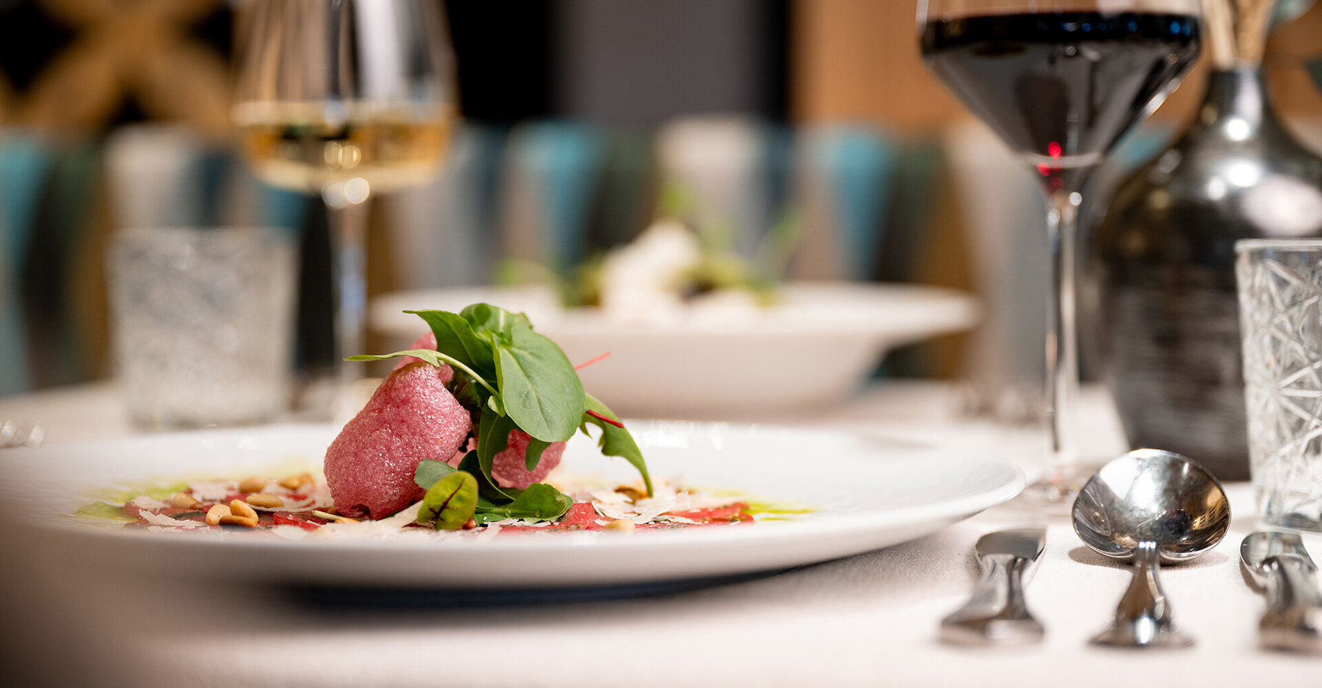 A close-up of a gourmet dish garnished with herbs, alongside glasses of red and white wine, on an elegantly set restaurant table under soft lighting.