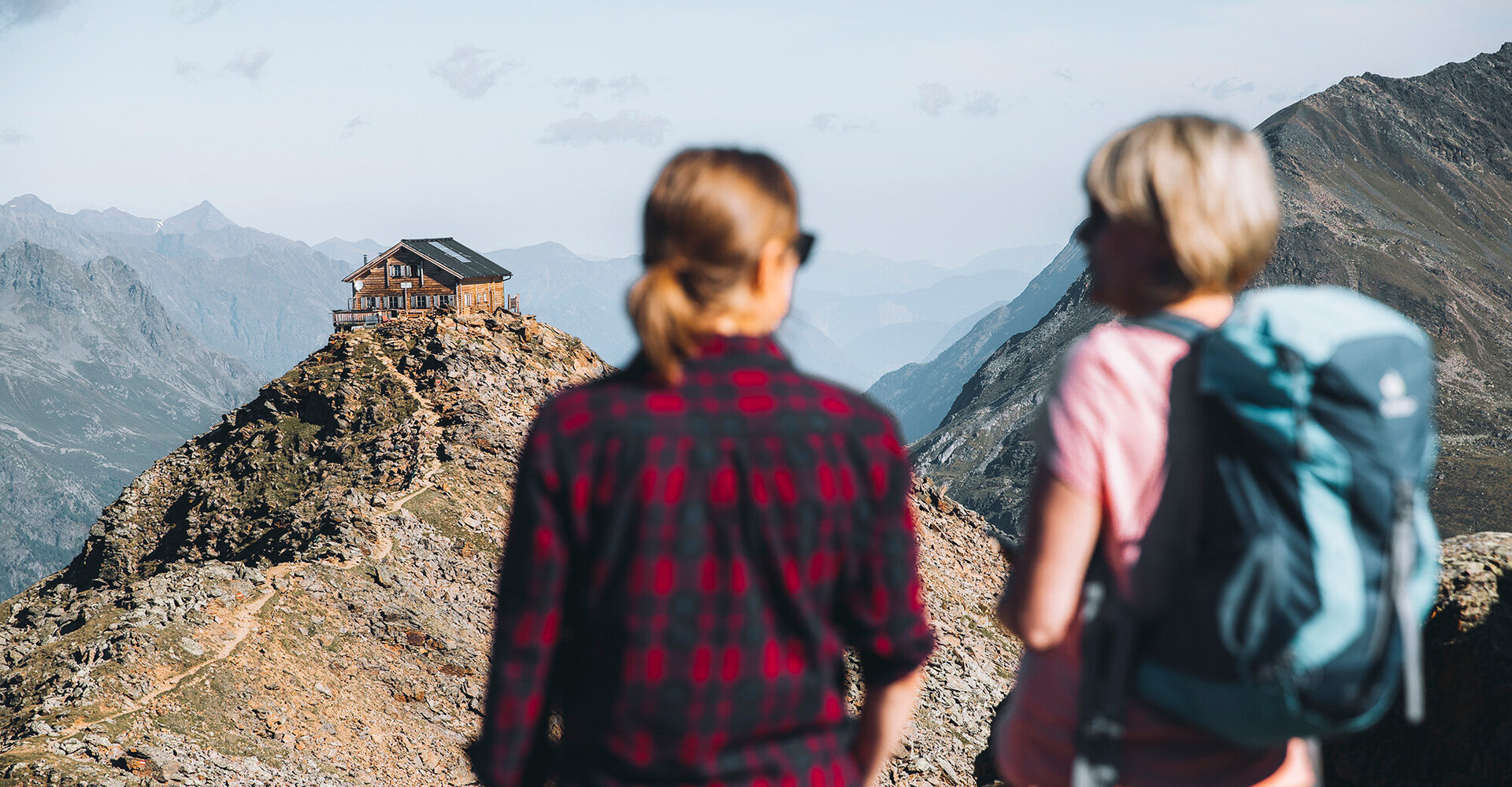 Zwei Wanderer stehen auf einem Bergpfad und blicken auf eine Hütte, die auf einem felsigen Bergrücken thront, umgeben von fernen Bergen unter einem klaren Himmel.