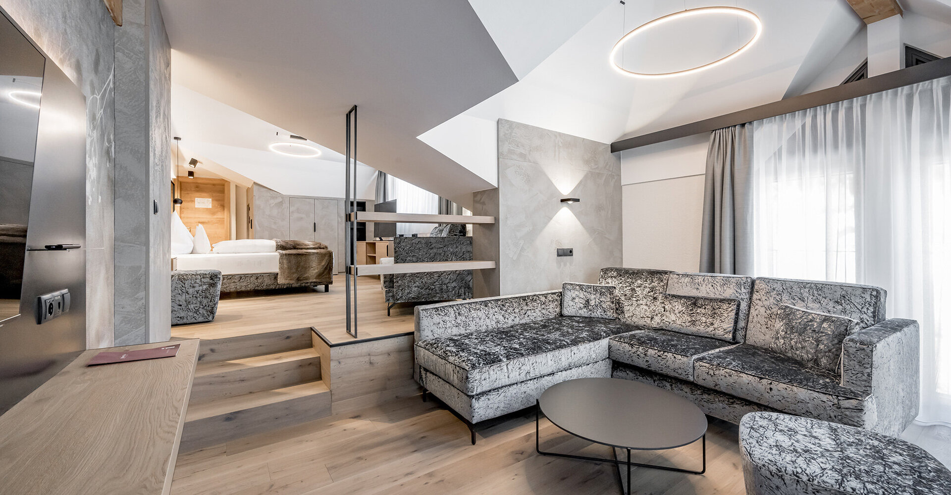 Moderne Suite im Loft-Stil mit einem bequemen grauen Ecksofa, einem runden Couchtisch und einem offenen Schlafbereich auf einer erhöhten Plattform. Große Fenster mit transparenten Vorhängen lassen viel Tageslicht herein.