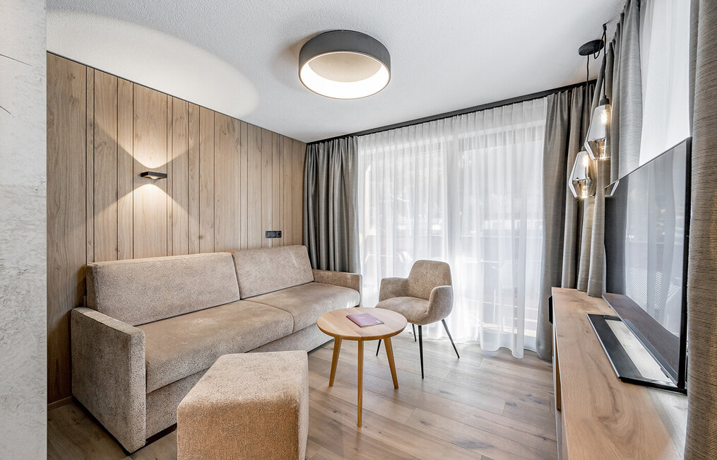 Modern living room with a beige sofa, an armchair, a round wooden table and a floor-to-ceiling window with sheer curtains. Neutral tones, wooden walls and a flat-screen TV create a cozy, minimalist atmosphere.