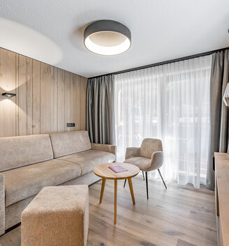 Modern living room with a beige sofa, an armchair, a round wooden table and a floor-to-ceiling window with sheer curtains. Neutral tones, wooden walls and a flat-screen TV create a cozy, minimalist atmosphere.