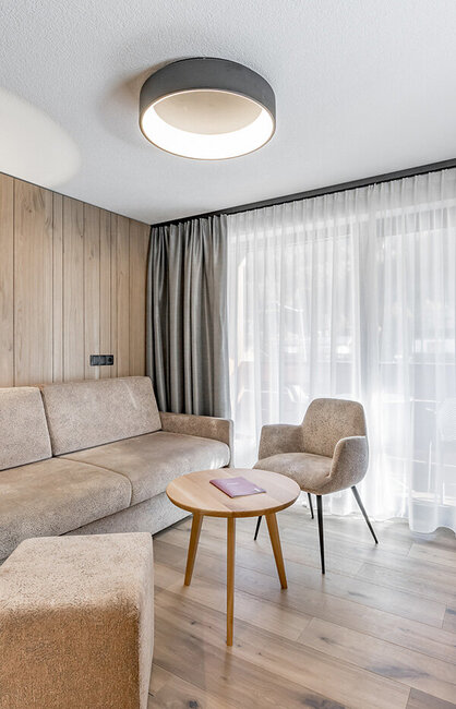 Modernes Wohnzimmer mit beigem Sofa, Sessel, rundem Holztisch und raumhohem Fenster mit transparenten Vorhängen. Neutrale Farbtöne, Holzwände und ein Flachbildfernseher schaffen eine gemütliche, minimalistische Atmosphäre.