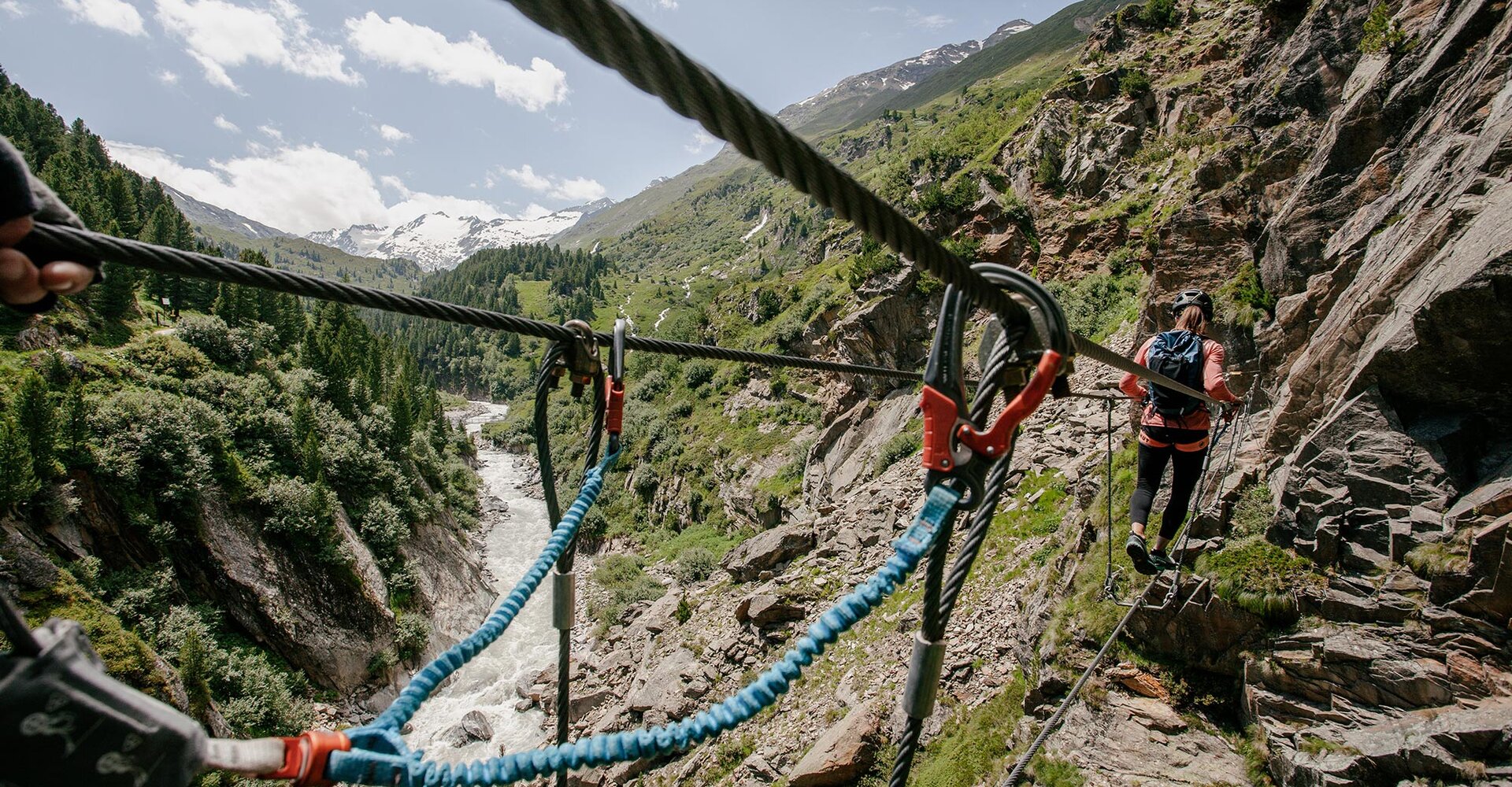 A person wearing a climbing harness traverses a high ropes course above a rocky valley with a river flowing below, surrounded by green trees and mountains under a partly cloudy sky.