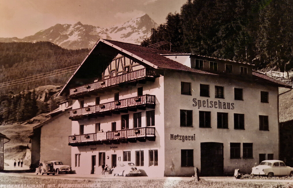 A historical photo of a building in the Alps bearing the inscription "Speisehaus Metzgerei" featuring balconies, cars parked in front, people nearby and wood-covered mountains in the background.