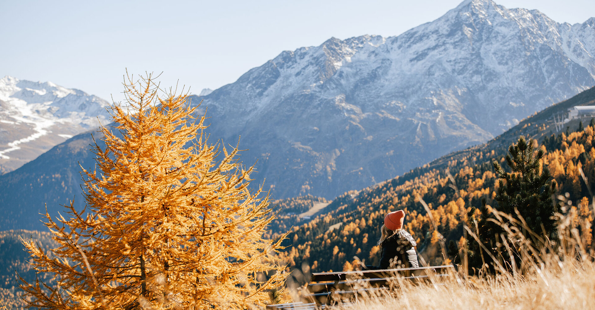 A person wearing a red hat sits on a bench, gazing under a clear sky at golden autumn trees and snow-covered mountains.