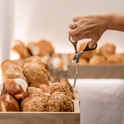 A hand using tongs to pick up a bread roll from a wooden tray filled with several rolls and pastries.