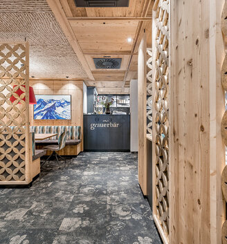 Modern restaurant interior featuring light wood paneling, decorative partition elements, carpeting and a bar area in the background with "Grauer Bär" lettering. A mountain painting and red lamps provide accents of color.