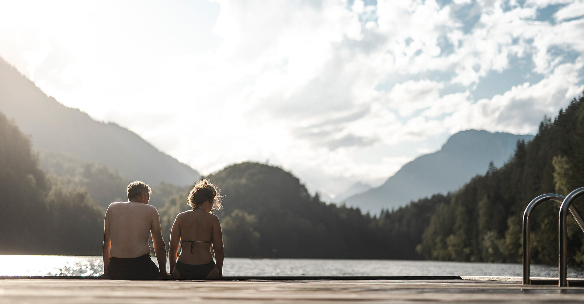 Zwei Menschen in Badekleidung sitzen auf einem Holzsteg an einem See, umgeben von bewaldeten Hügeln und Bergen unter einem teilweise bewölkten Himmel. Neben ihnen führt eine Metallleiter ins Wasser hinab.