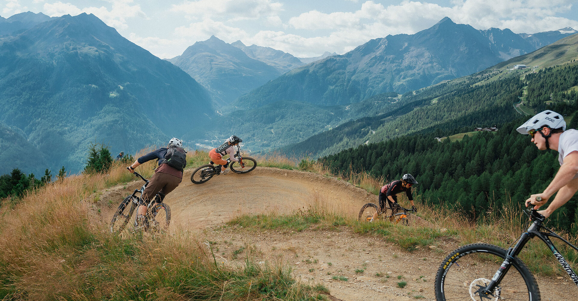 Four mountain bikers ride down a trail on a slope, surrounded by green trees and high mountains under a partly cloudy sky.