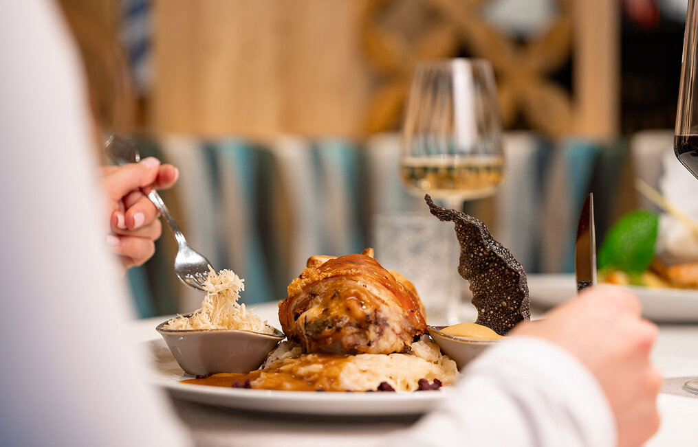A person at a restaurant table is holding a fork and knife and enjoying a gourmet meal, featuring meat, sauce and side dishes. Two wine glasses are in the background. 