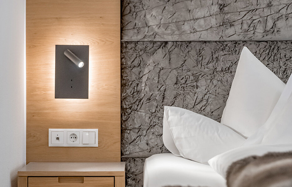 A modern hotel room featuring a wooden nightstand, a textured gray headboard, white bedding and a small wall-mounted reading lamp. A fluffy blanket lies across the bed.
