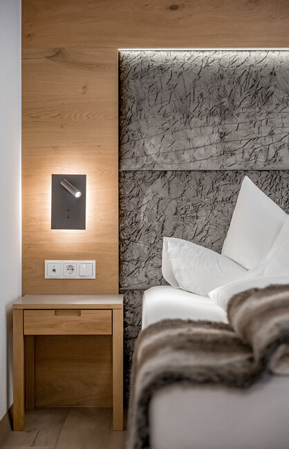 Ein modernes Hotelzimmer mit einem Nachttisch aus Holz, einem grauen Kopfteil mit Struktur, weißer Bettwäsche und einer kleinen, an der Wand montierten Leselampe. Über dem Bett liegt eine flauschige Decke.