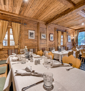 Cozy wooden restaurant interior featuring wooden tables, upholstered benches and natural light streaming in through large windows. Framed photos hang on the walls, creating a warm, rustic atmosphere.
