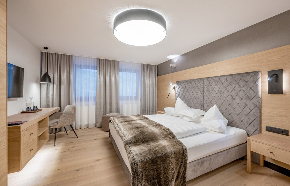 A modern hotel room featuring a large bed, a faux-fur throw, a desk, a chair, a wall-mounted TV and a large ceiling light. The décor uses neutral tones with accents of wood and upholstered furniture. Two windows with sheer curtains allow daylight to stream in.