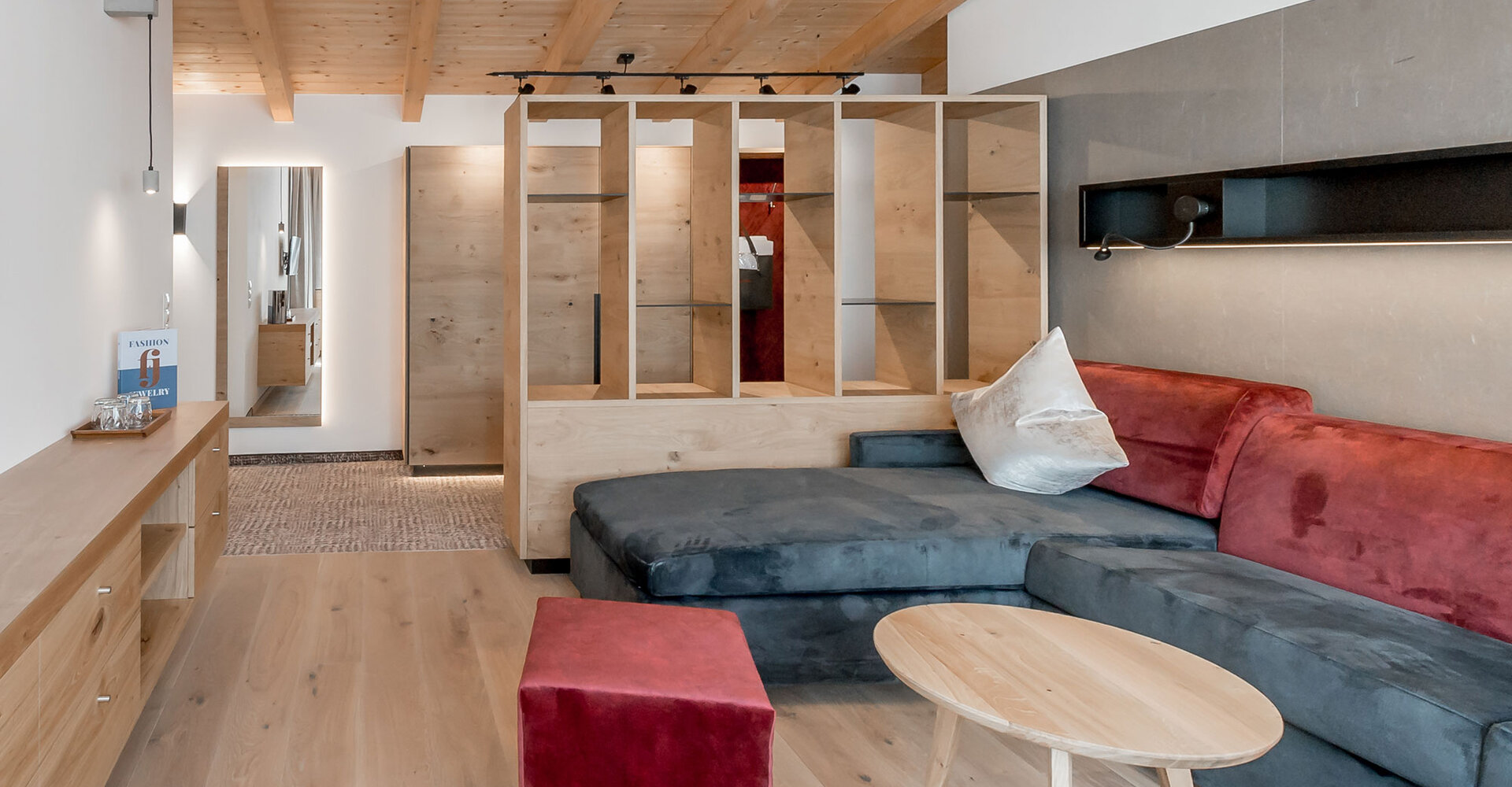 Modern living room with wooden flooring, an L-shaped sofa in gray and red, a small round wooden coffee table, a wooden room divider and a view into a hallway with a mirror hanging at the far end. Exposed wooden beams are on the ceiling.