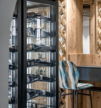 In a modern, stylish interior, a glass-fronted wine cooler filled with wine bottles stands next to a striped bar stool and wood paneling.