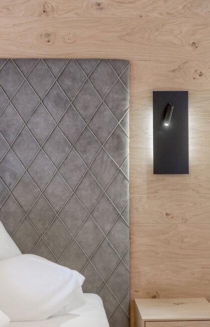 Ein graues Kopfteil mit Rautenmuster neben einer Holzwand mit einer modernen schwarzen Wandleuchte und einem kleinen Nachttisch aus Holz, auf dem ein weißes Kissen teilweise zu sehen ist.