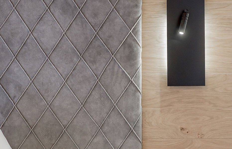 A gray headboard with a diamond pattern next to a wooden wall featuring a modern black wall lamp and a small wooden nightstand with a white pillow.