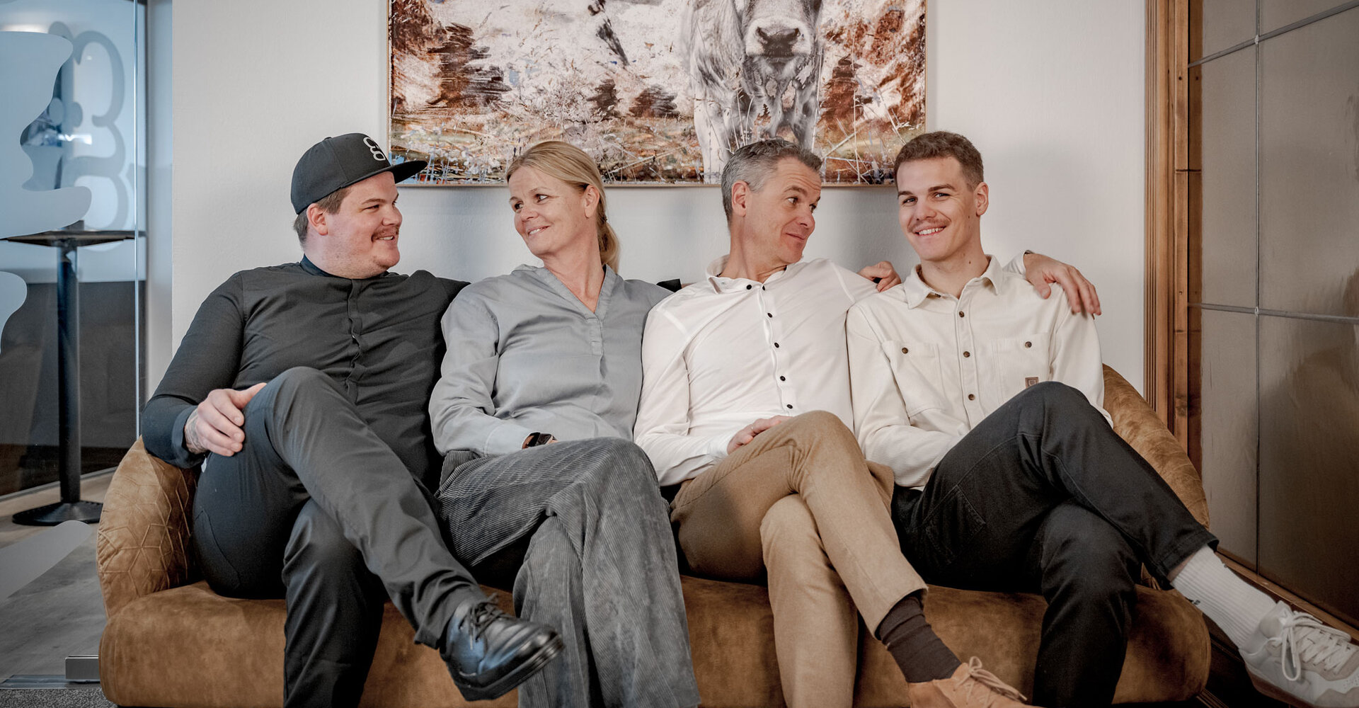 The Gstrein family sits close together on a brown sofa, smiling and looking at each other. On the wall behind there is a painting of a cow, the entire setting appears to be a modern seating area.