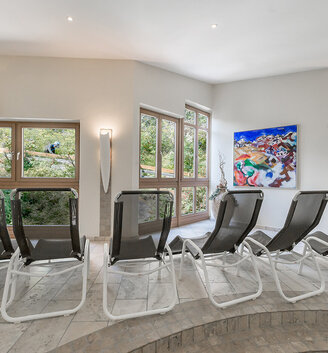 A bright relaxation room with black loungers, large windows overlooking the greenery, a colorful painting on the wall and a marble floor.
