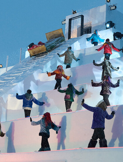 People in colorful winter clothing stand spaced apart on icy steps and perform a synchronized dance sequence outdoors on a snow structure, while lights illuminate the scene.
